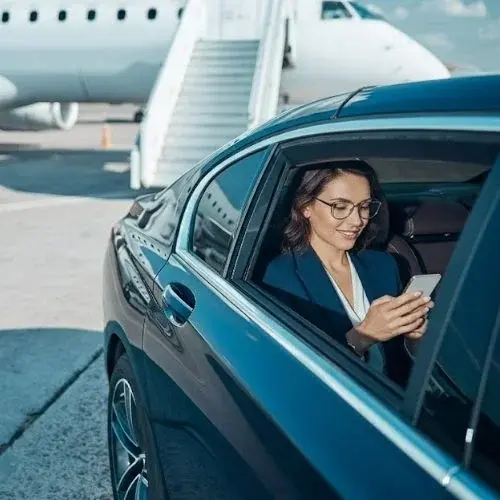 A female professional in a blue suit smiling and using a phone in the back seat of a black car with a plane and stairs in the background A female professional in a blue suit smiling and using a phone in the back seat of a black car with a plane and stairs in the background