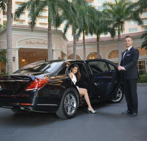 A professional chauffeur opens the door of a luxurious black sedan for a woman in a white dress at a tropical resort entrance A professional chauffeur opens the door of a luxurious black sedan for a woman in a white dress at a tropical resort entrance