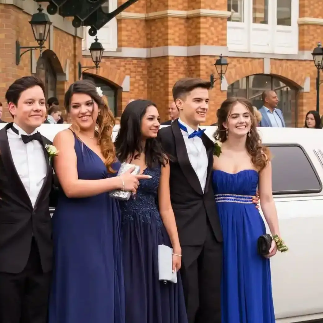 Five teenagers in formal prom attire smiling and posing in front of a white luxury limousine